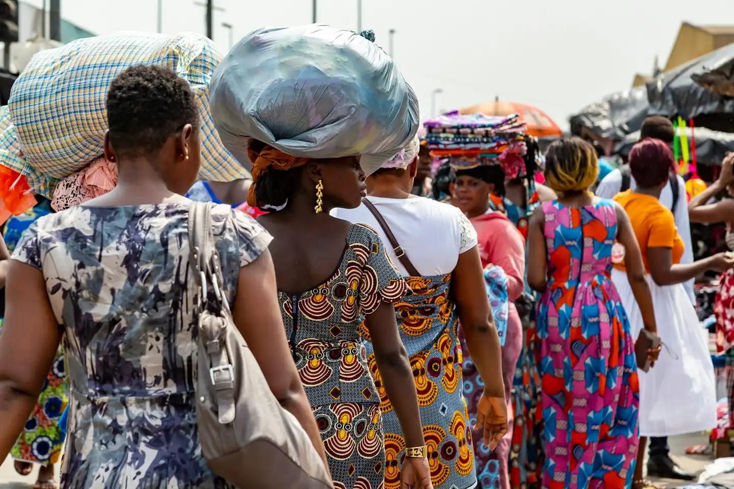 Des femmes, nombre d’entre elles en robes de pagne imprimé, traversent une rue de marché animée, plusieurs portant de gros ballots sur la tête.