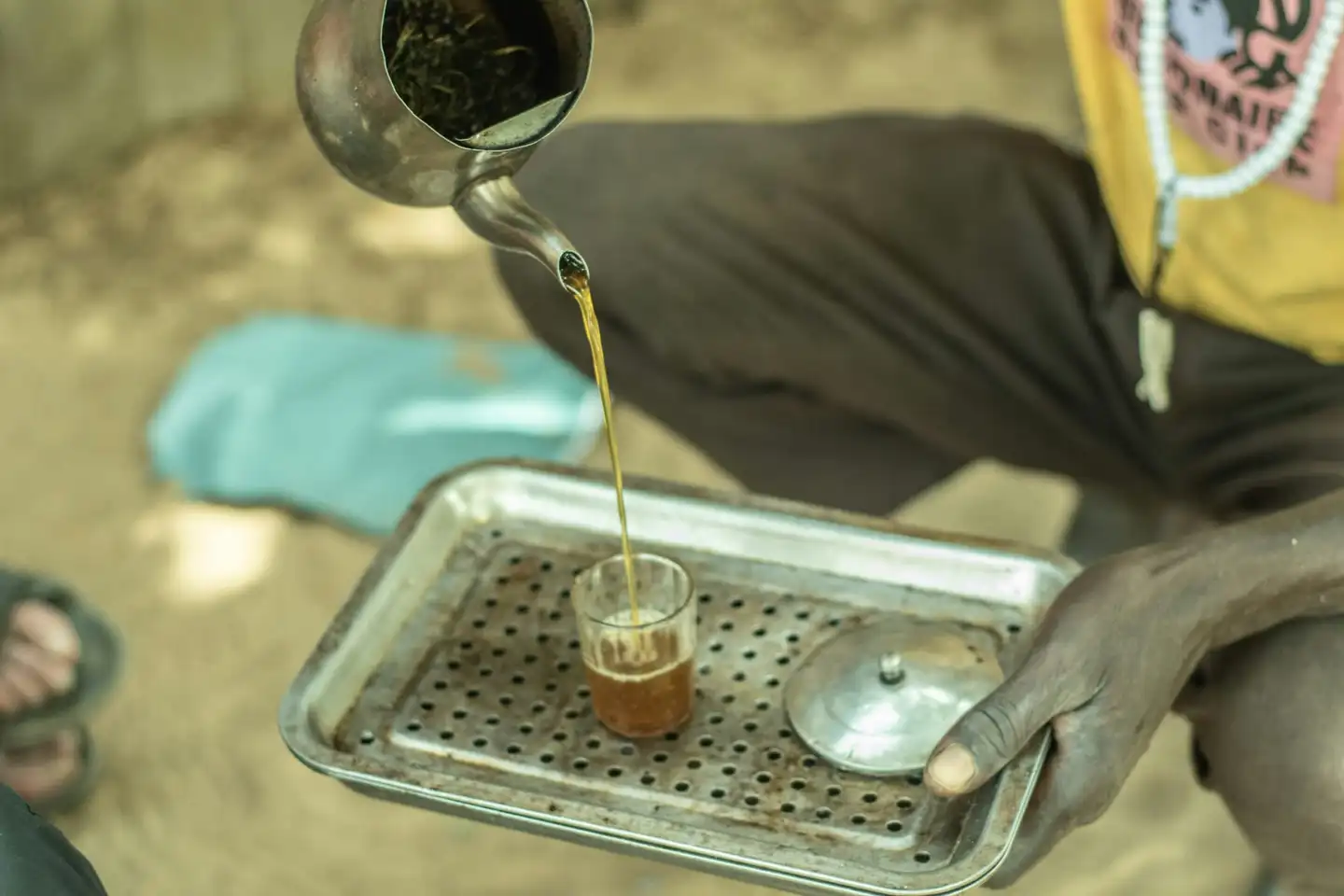 A person pours a thin stream of tea from a small metal teapot into a glass on a perforated metal tray.