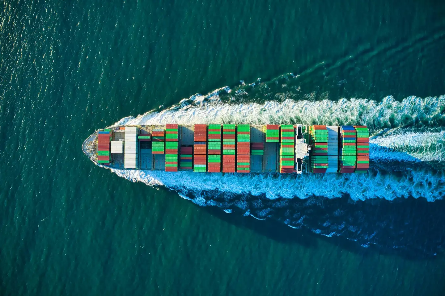 Top-down aerial view of a container ship stacked with red and green containers, leaving a white wake on deep green water.