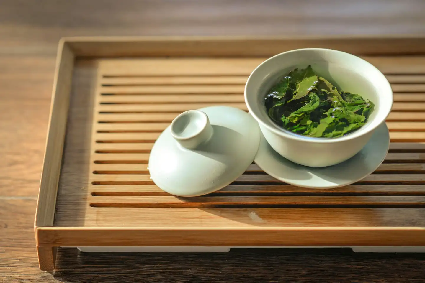 A pale porcelain lidded bowl of steeped green tea leaves on a slatted wooden tea tray, its lid resting beside it.