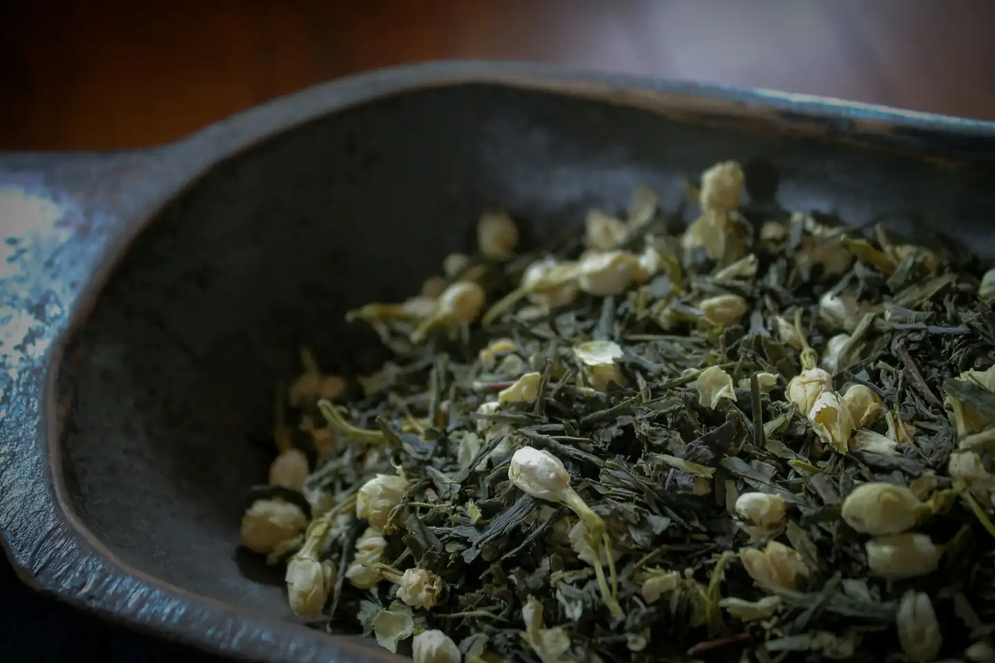 Dry green tea leaves mixed with small cream-white dried flower buds, in a dark, worn scoop.