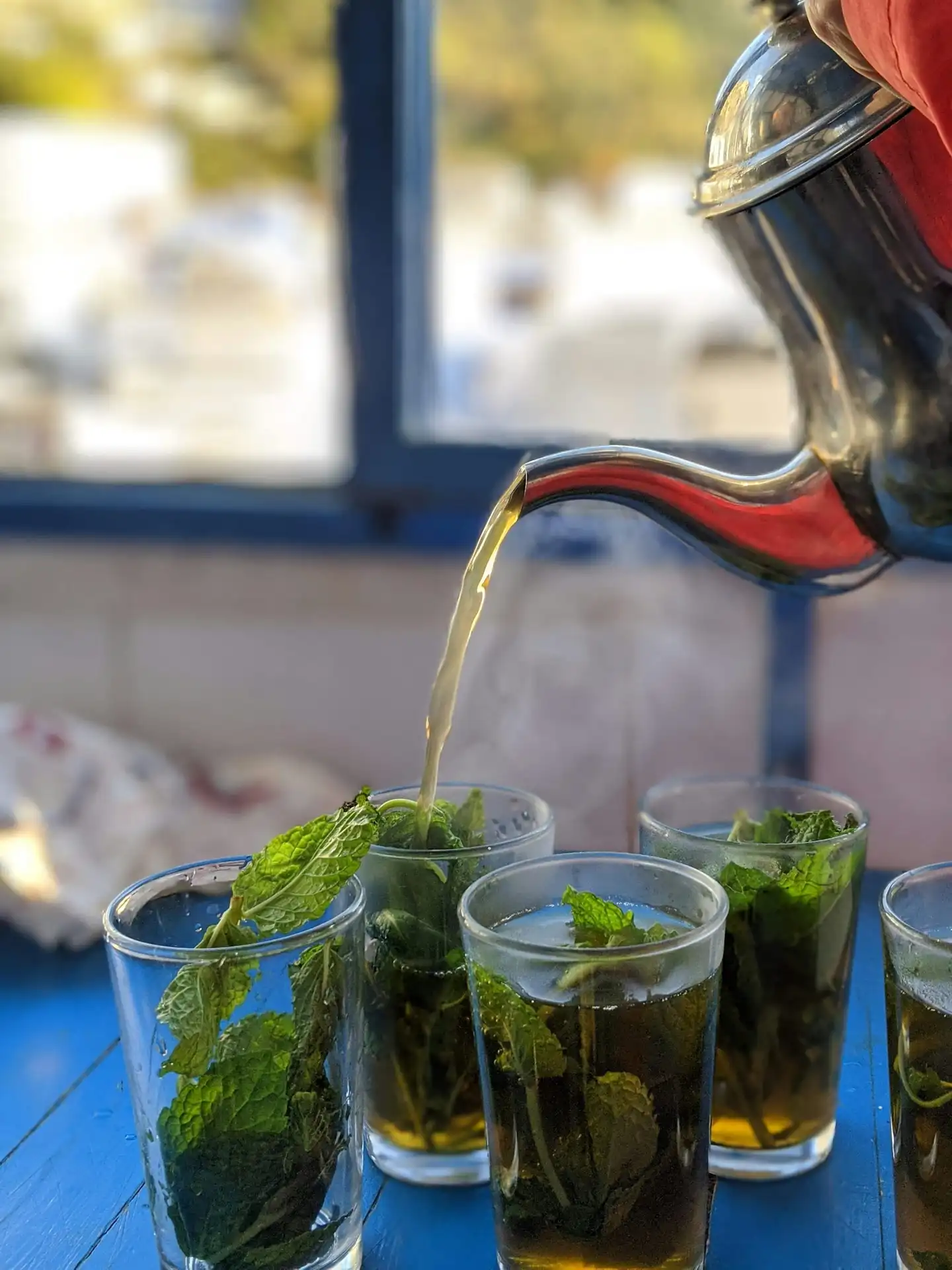 Mint tea poured from a silver teapot into glasses packed with fresh mint leaves, on a blue table.